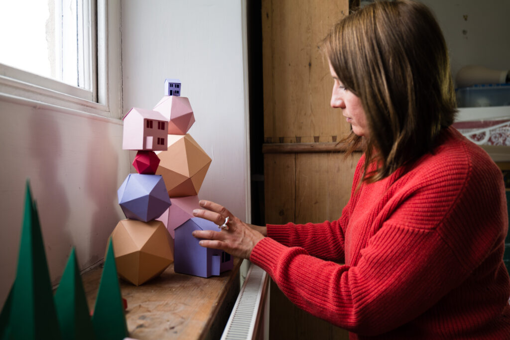 Photo shows Amy Williams, a brunette woman in a red jumper, balancing colourful spheres and houses made of paper.