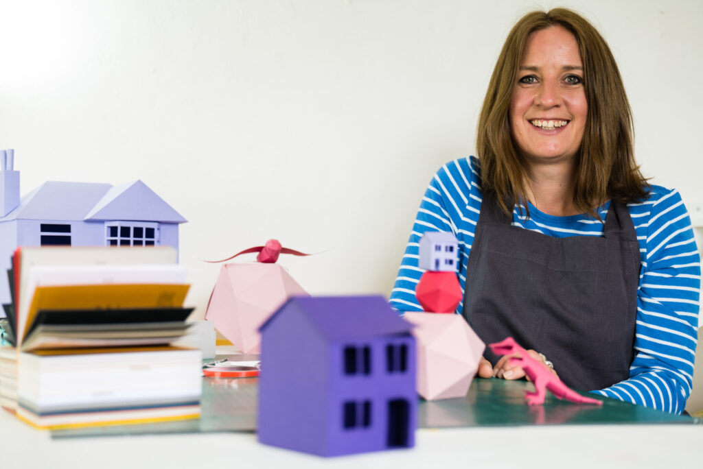 The photo shows Amy Williams, a brunette woman with shoulder-length brown hair. She is smiling. On the table in front of her are 3D creations made from paper, including purple houses, a small pink dinosaur and geometric pink spheres.