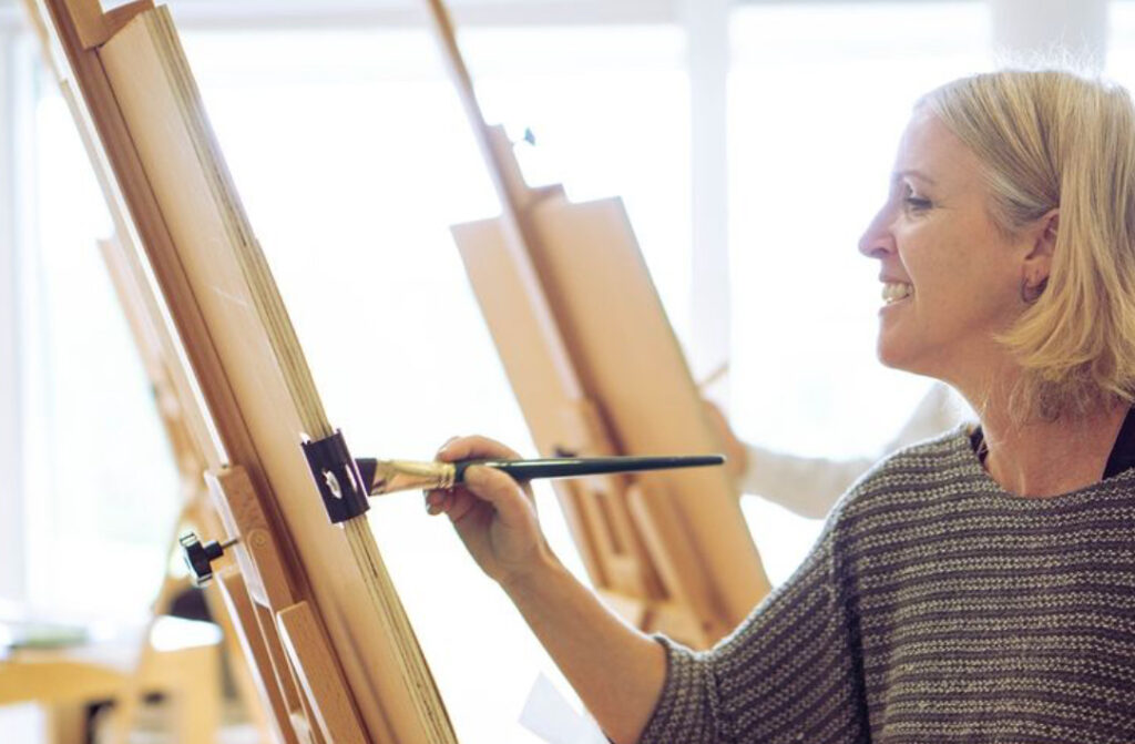 Photo shows Catherine MacDiarmid, a woman with blonde shoulder length hair, holding a brush to a canvas which is facing side on.
