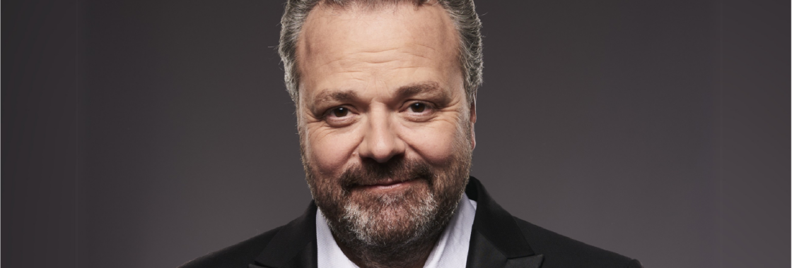Hal Cruttenden against a black backdrop smiling to camera.