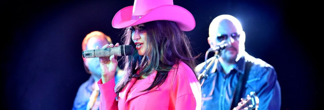 Shania tribute act wearing a pink cowboy hat and a pink blazer with black hair. Behind her are two men - one playing the guitar, the other obscured by Shania