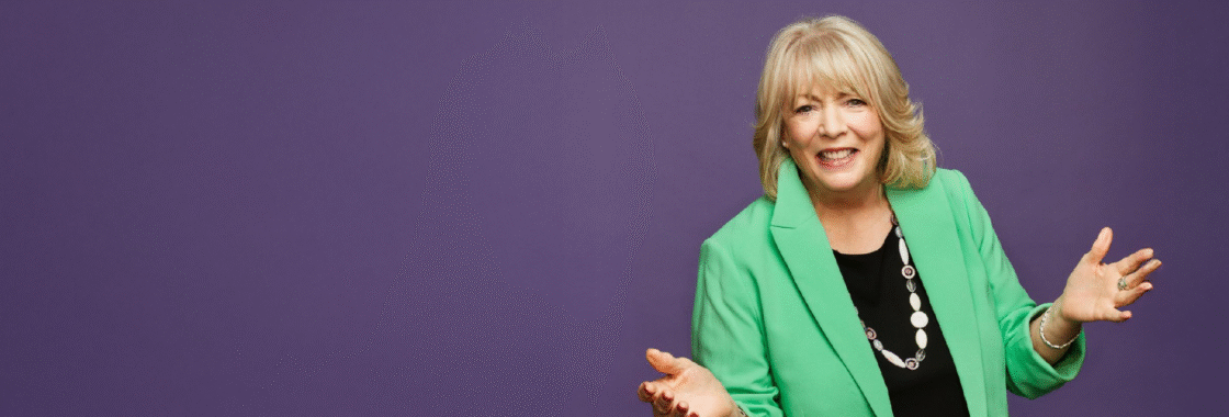 Alison Steadman wearing a green blazer, arms outstretched against a purple background